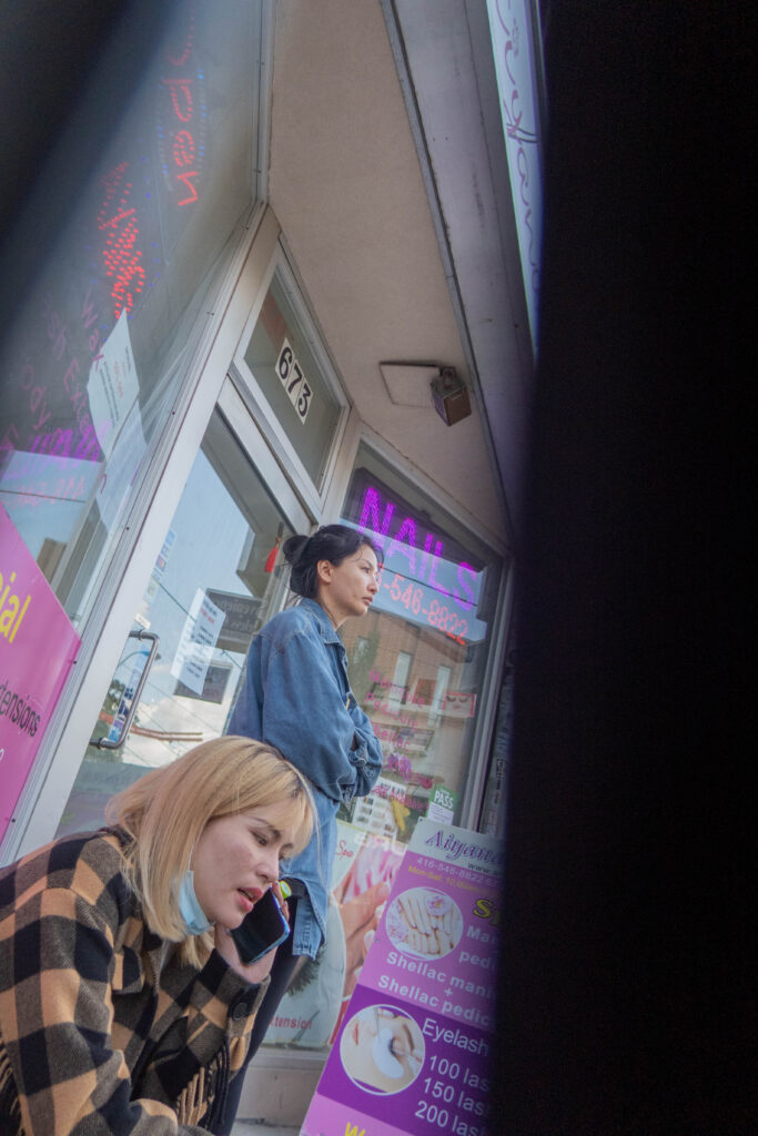 Two women sitting outside a nail salon, one looking into the distance as the other talks on the phone. 