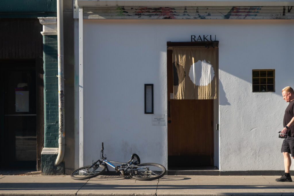 Store front of Raku restaurant in Toronto with a bicycle and a man in all black outside on the sidewalk. 